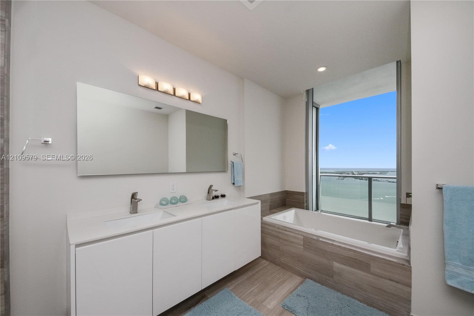 Double vanity with integrated sinks, frameless wall-to-wall mirror, built-in soaking tub with stone-finish surround, wood-finish flooring, and floor-to-ceiling windows