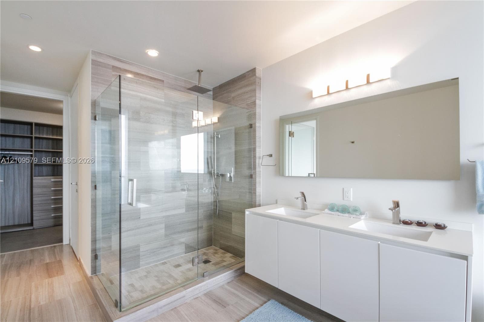 Dual-sink vanity with white cabinetry and rectangular basins, oversized frameless vanity mirror, glass-enclosed walk-in shower with tiled walls and rainfall showerhead, and integrated wardrobe cabinetry