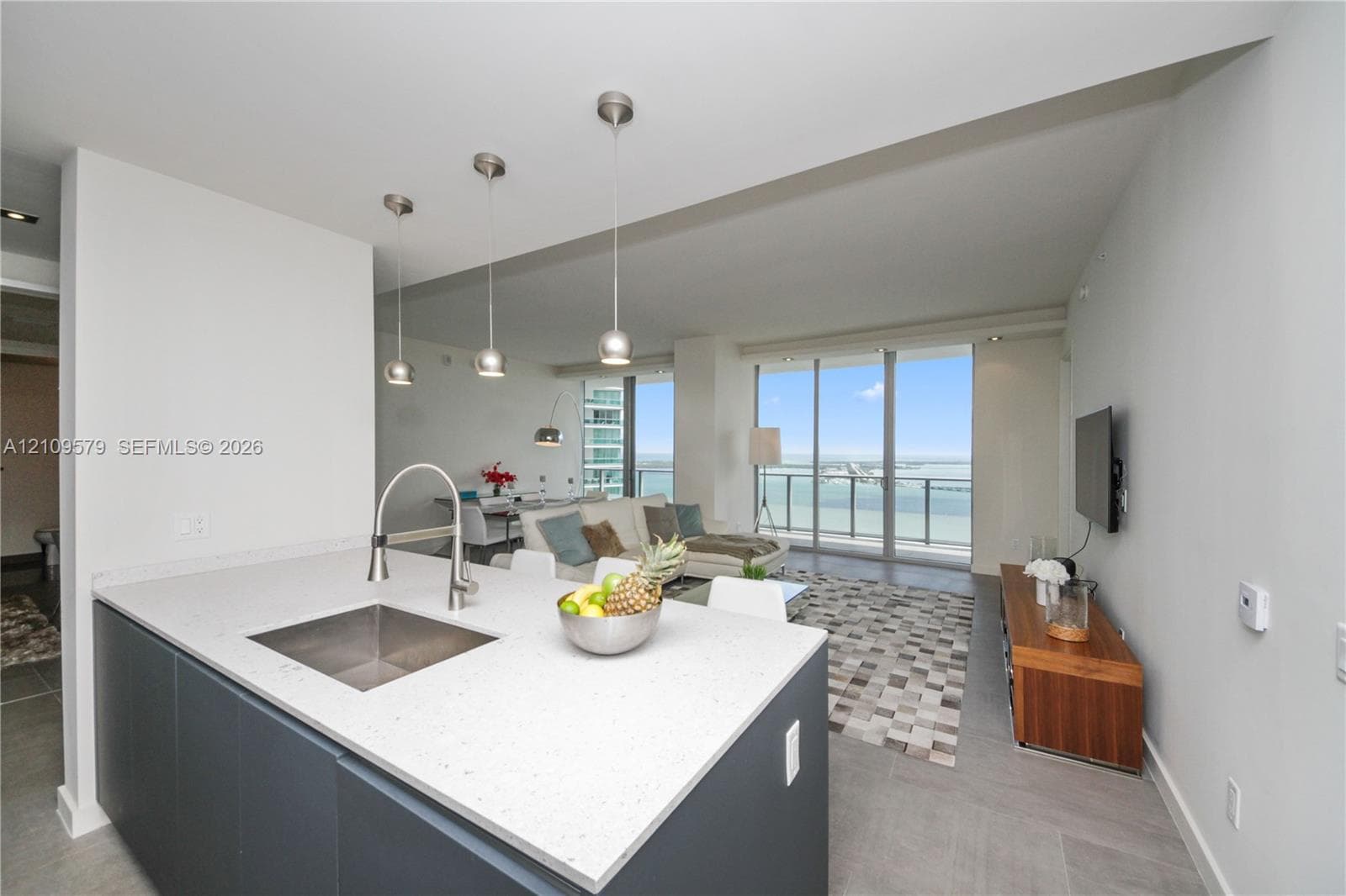 Kitchen island featuring stone countertops, a stainless steel under-mount sink, matte-finish cabinetry, and three pendant light fixtures, with expansive floor-to-ceiling glass sliding doors opening to a balcony
