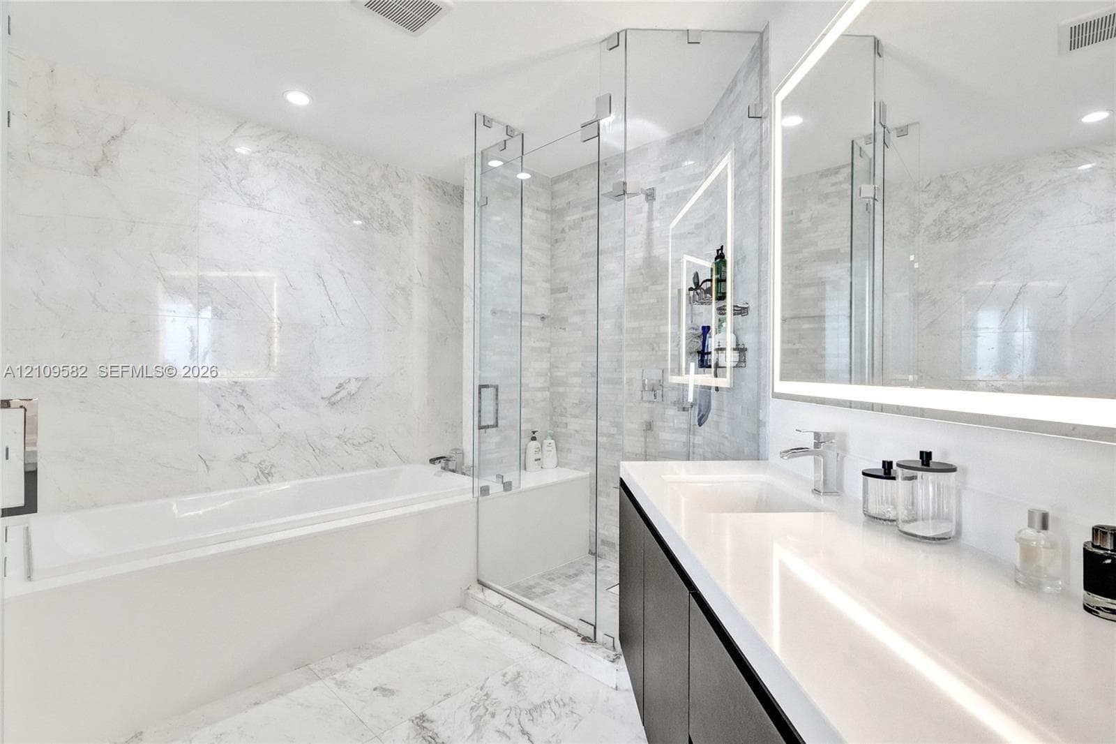 Bathroom featuring a deep soaking tub, glass-enclosed shower with tiled walls, stone-finish flooring, dual-sink vanity with solid-surface countertop, and an integrated LED-lighted mirror
