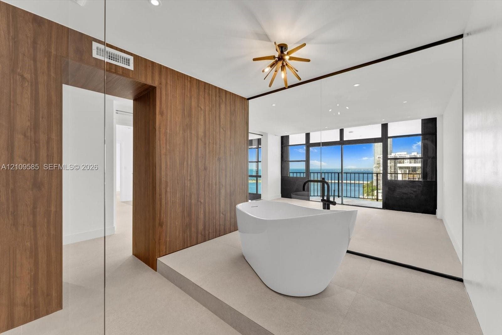 Freestanding soaking tub with matte black faucet fixture, elevated platform, wood-finish wall paneling, multi-light geometric chandelier, and floor-to-ceiling windows