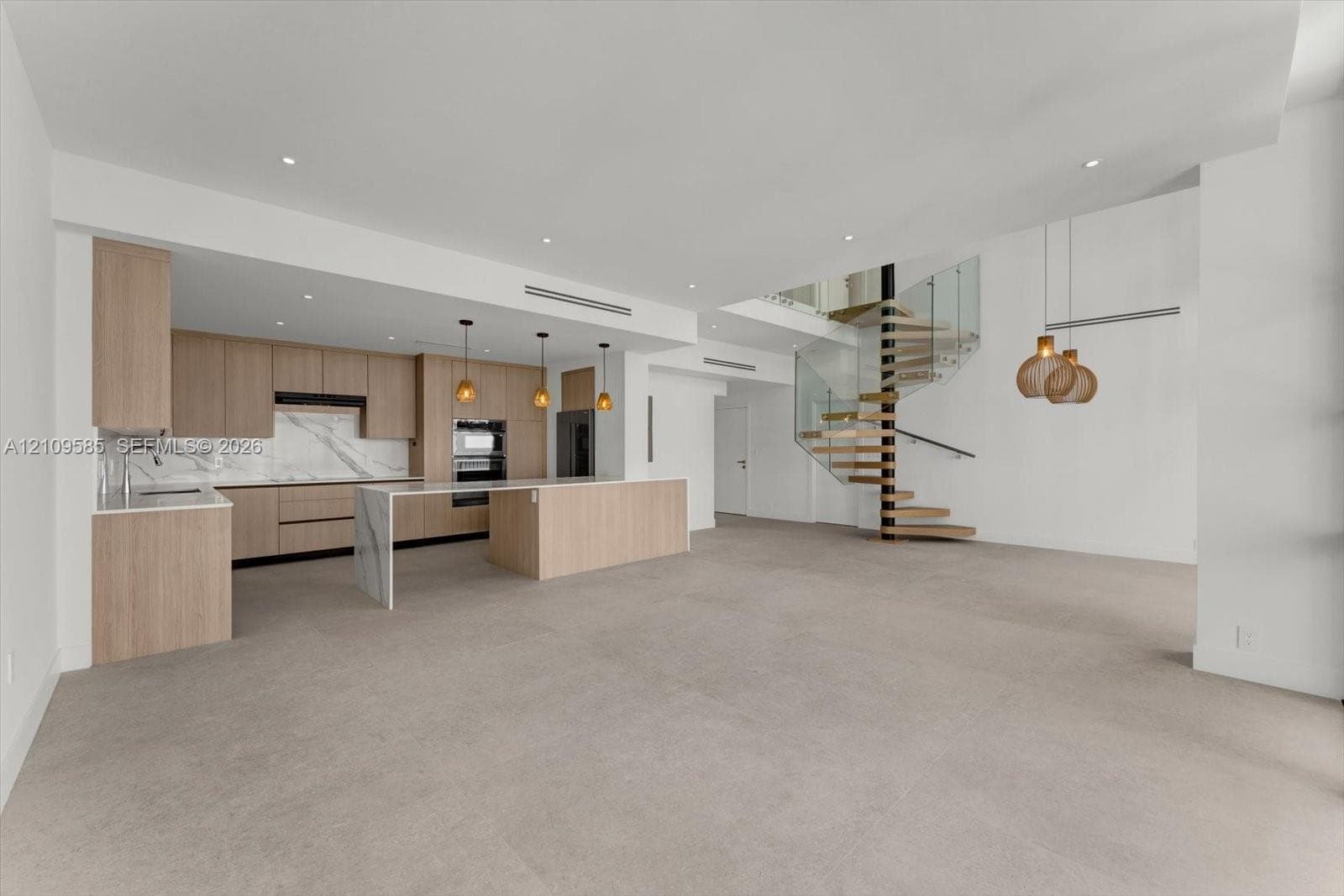 Open-concept interior featuring light wood-finish cabinetry, waterfall-edge stone island, spiral staircase with glass railing, and recessed ceiling lighting