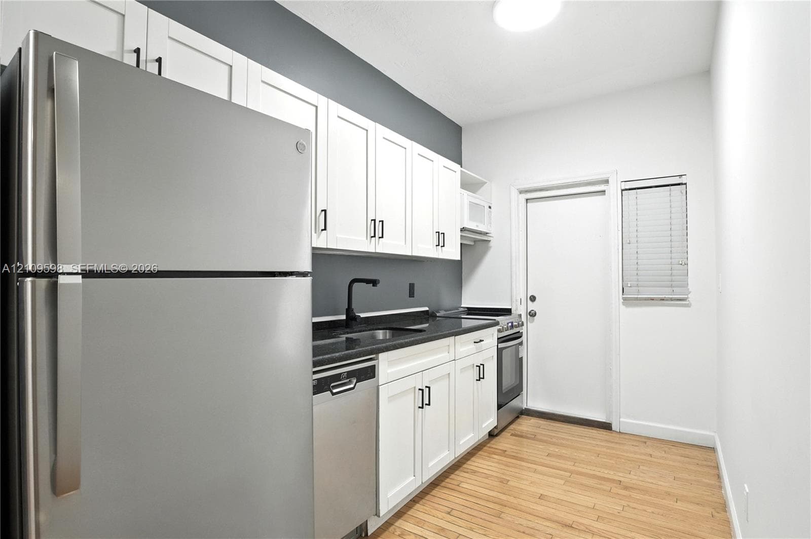 Kitchen featuring white shaker-style cabinetry, dark stone-finish countertops, stainless steel appliances, a matte black faucet, and wood-finish flooring