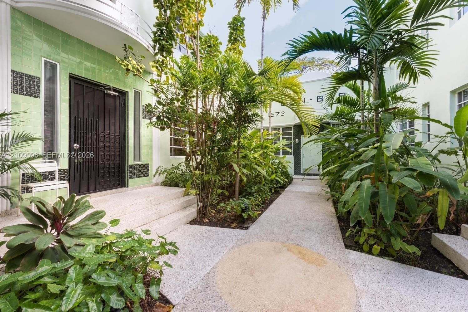 Art Deco exterior featuring light green decorative tile facade, terrazzo pathways, dark metal entryway doors, and lush tropical landscaping