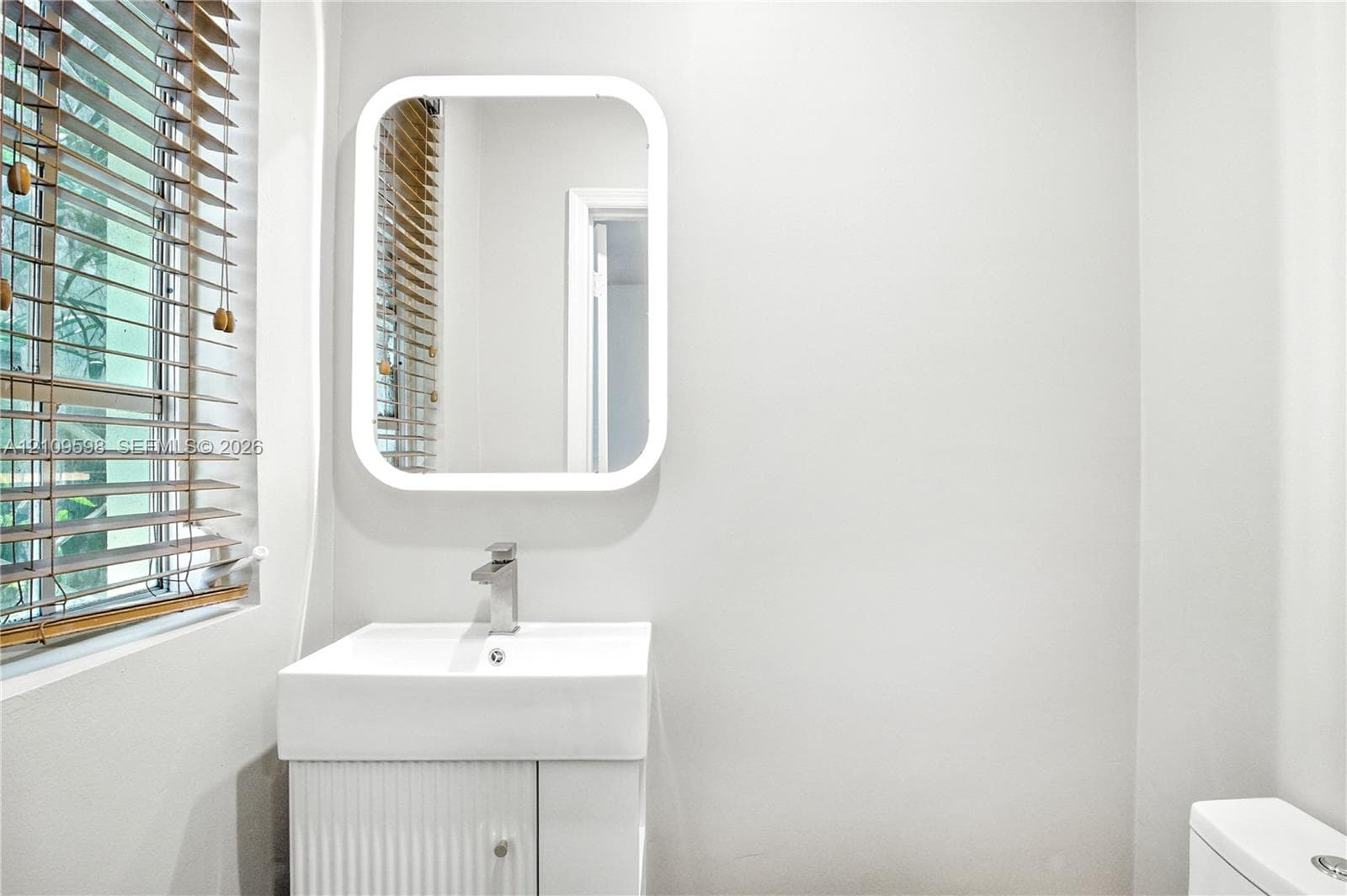 Wall-mounted rectangular mirror with integrated LED perimeter lighting, white ribbed vanity cabinet, rectangular basin, polished chrome faucet, and horizontal slatted window blinds