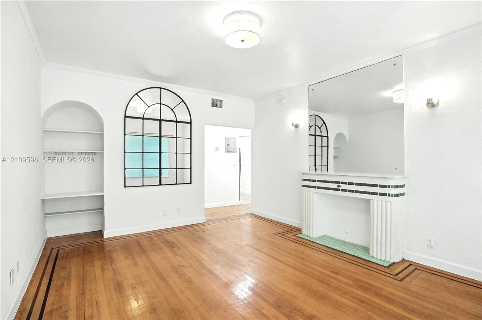 Living area featuring wood-finish flooring with inlay detailing, a fireplace mantle with fluted columns, a decorative archway window, a recessed arched alcove with built-in shelving, and crown molding