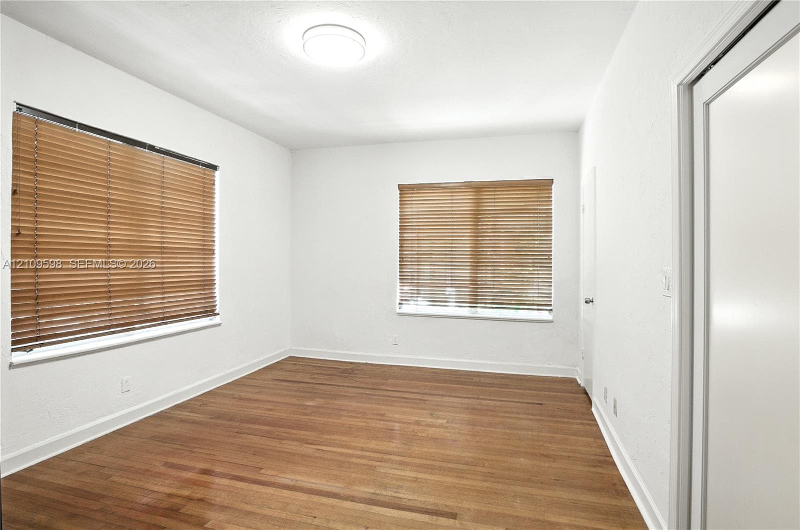 Interior room featuring wood-finish flooring, white walls, dual windows with horizontal blinds, and a ceiling-mounted light fixture