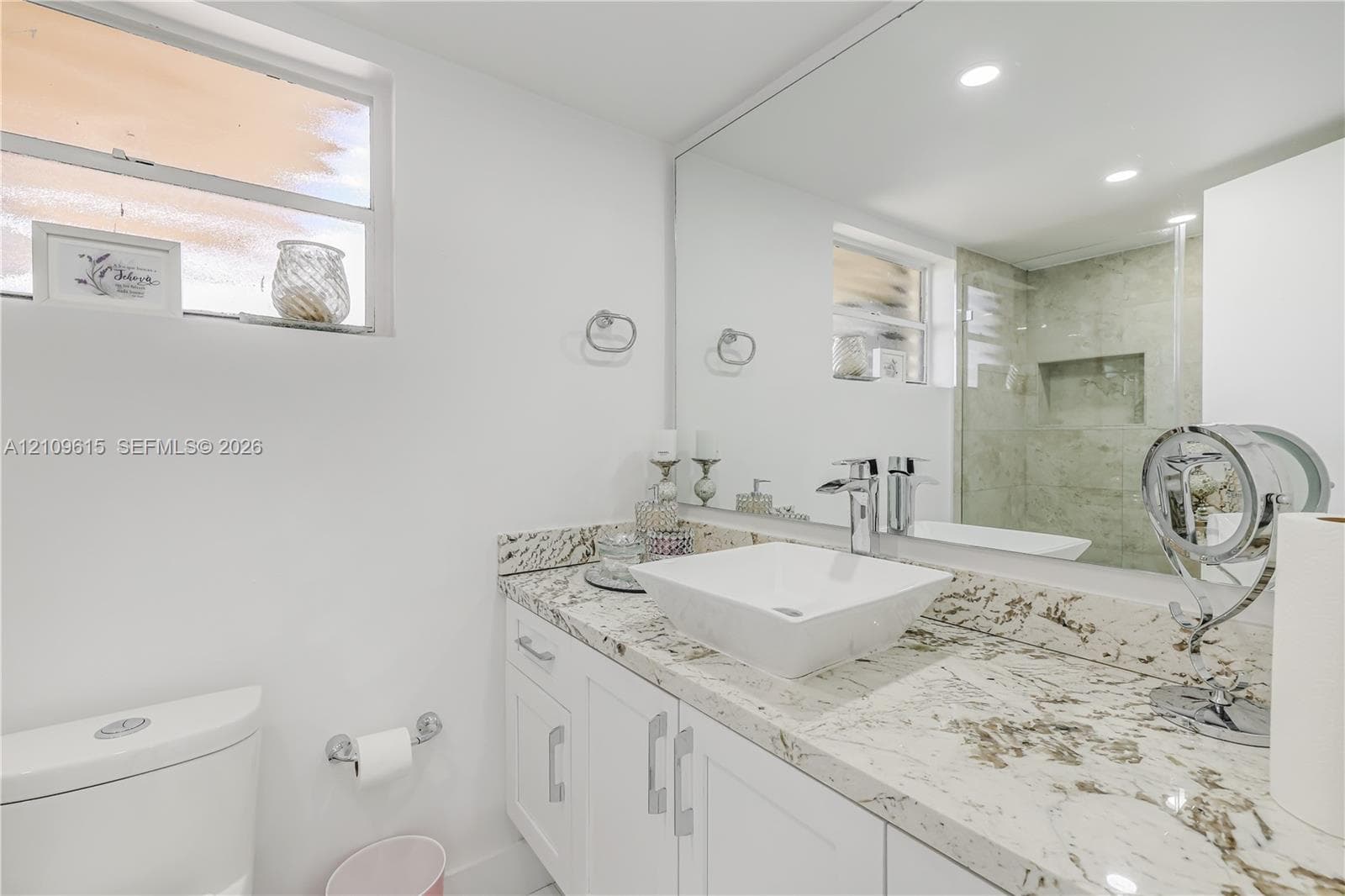 Bathroom featuring a white vanity cabinet, light-toned stone countertop, square vessel sink, chrome faucet, and glass-enclosed shower with a recessed wall niche