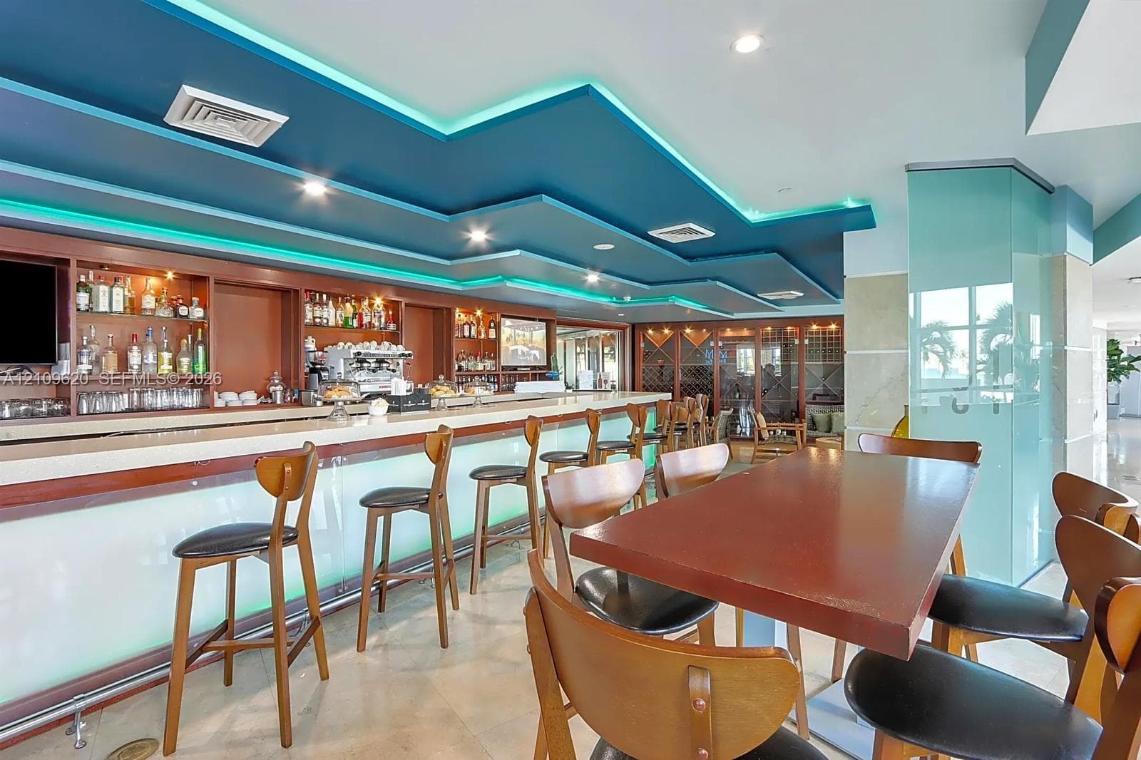Custom bar featuring a stone-topped counter, illuminated glass paneling, built-in wooden shelving, integrated wine storage, and a tiered drop ceiling with recessed lighting