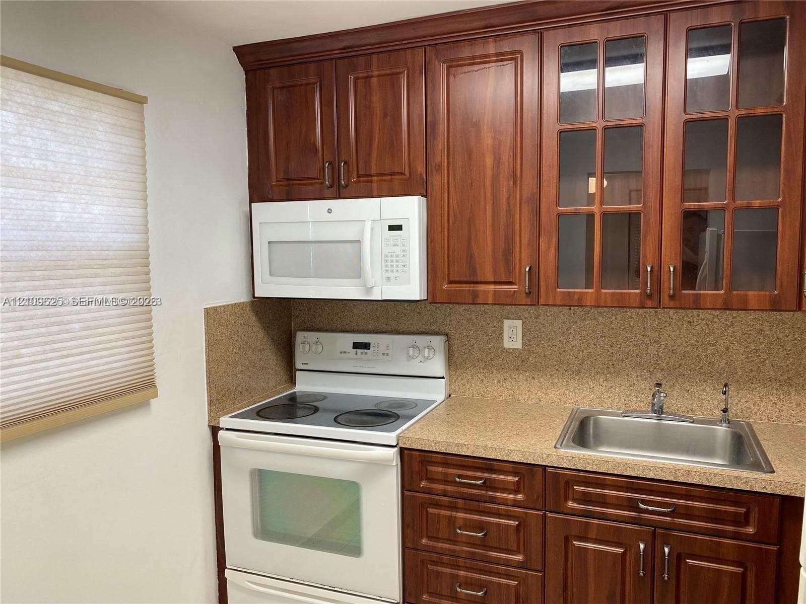 Kitchen featuring dark wood-finish cabinetry, glass-paneled upper cabinets, a textured countertop, a stainless steel sink, and white appliances