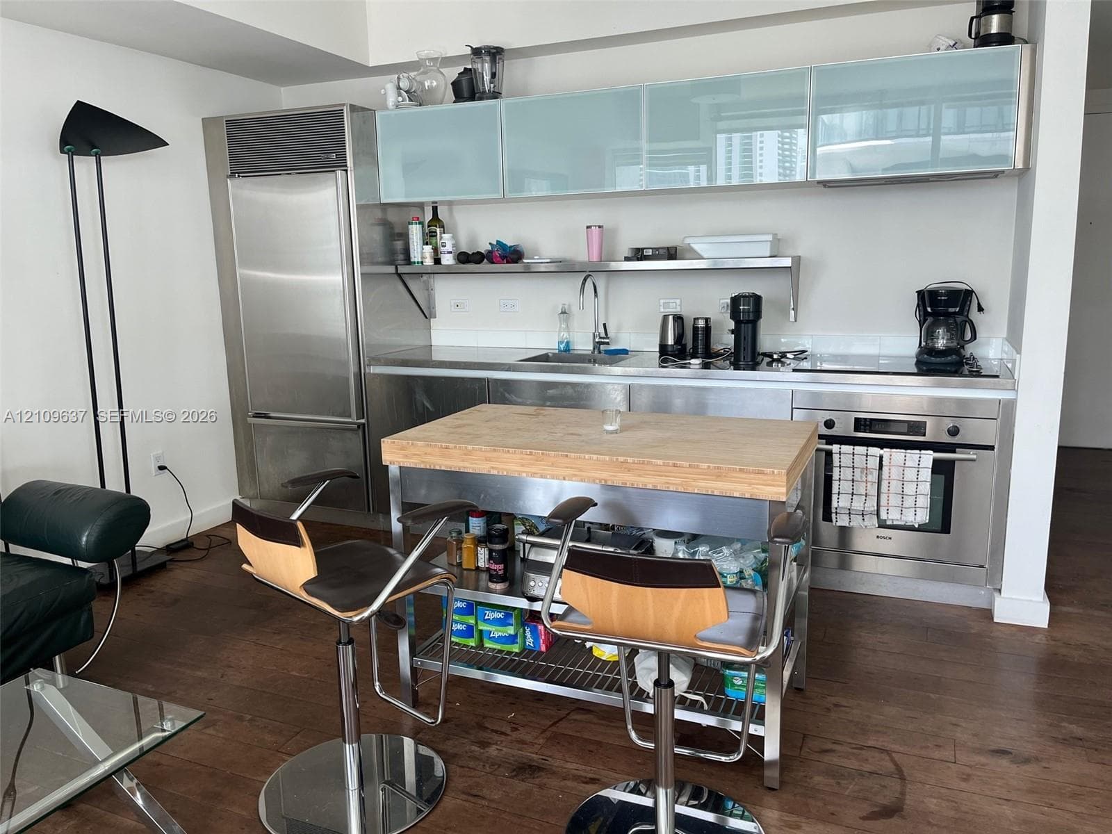 Stainless steel kitchen featuring a built-in refrigerator, under-cabinet lighting, frosted glass upper cabinetry, wood-finish flooring, and a sleek surface-mounted cooktop