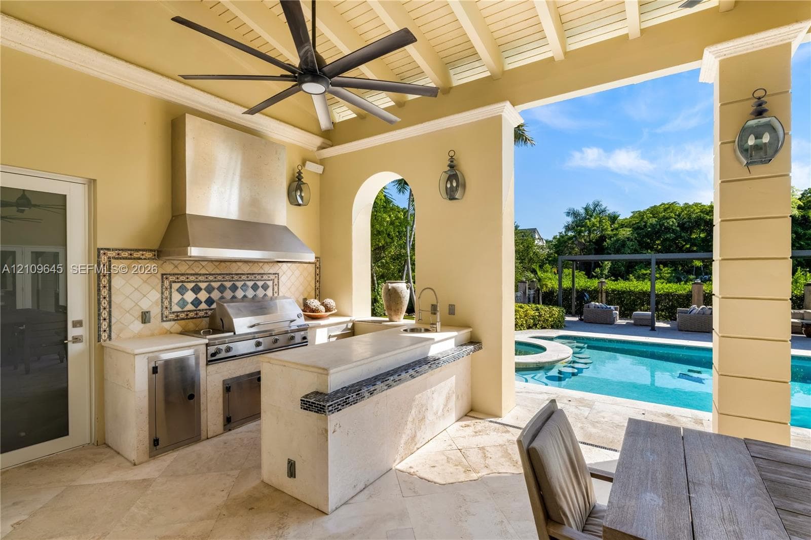 Covered outdoor kitchen featuring stone countertops, a stainless steel grill, a tile backsplash, and a built-in sink, accented by an arched opening and a pool with integrated seating