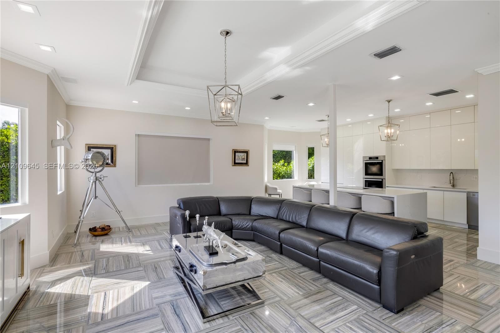 Open-concept area featuring high-gloss cabinetry, stone tile flooring, tray ceilings, recessed lighting, and a kitchen island with integrated sink