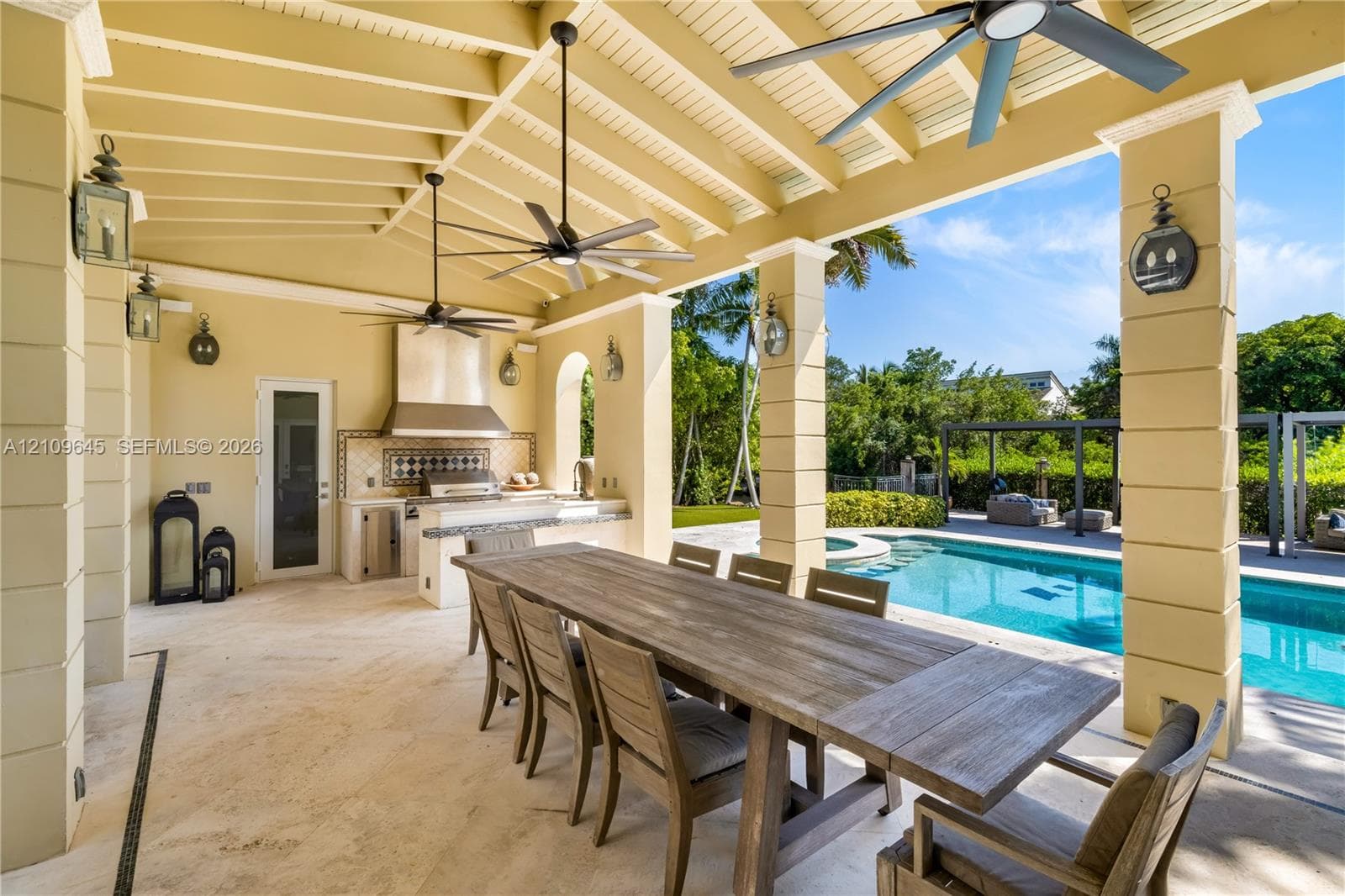 Covered outdoor patio featuring stone flooring, vaulted beamed ceiling with multiple ceiling fans, built-in kitchen with stainless steel range hood, and tile backsplash
