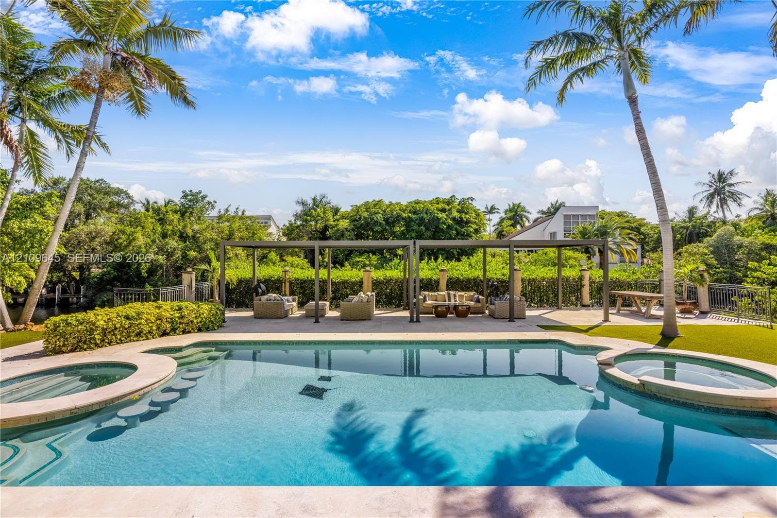 In-ground swimming pool with built-in wading stools and integrated spa, complemented by a stone-paved pool deck, covered pergola structure, and mature palm trees