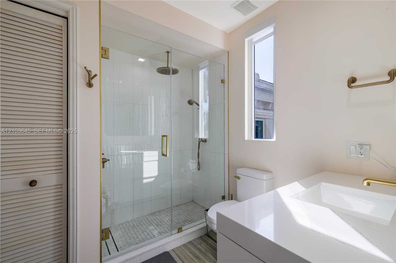 Walk-in glass shower featuring a rain-style showerhead and handheld sprayer with brass hardware, complemented by a white vanity with a rectangular integrated sink and a louvered-door closet