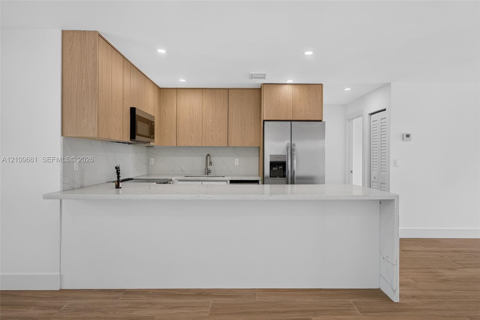 Kitchen featuring light wood-finish cabinetry, white stone-finish countertops, waterfall-edge island, integrated stainless steel appliances, and wood-finish flooring