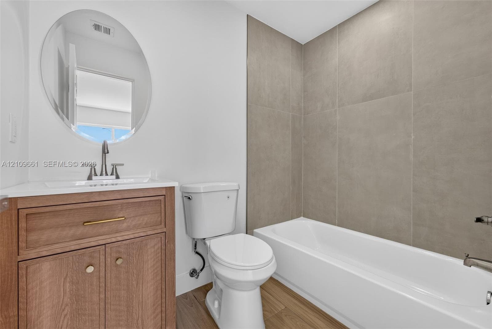 Bathroom featuring a vanity with wood-finish cabinetry, a white countertop, an oval wall-mounted mirror, and a tub with a floor-to-ceiling tile surround