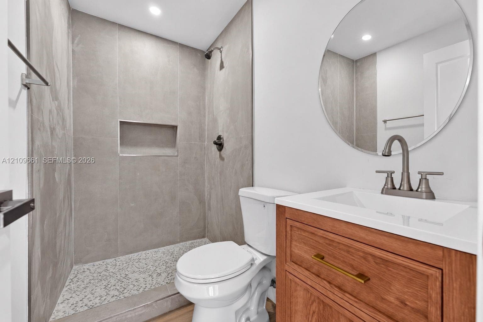 Walk-in shower with large-format grey wall tile and a built-in recessed niche, complemented by a wood-finish vanity with a white countertop, metallic hardware, and a circular wall-mounted mirror