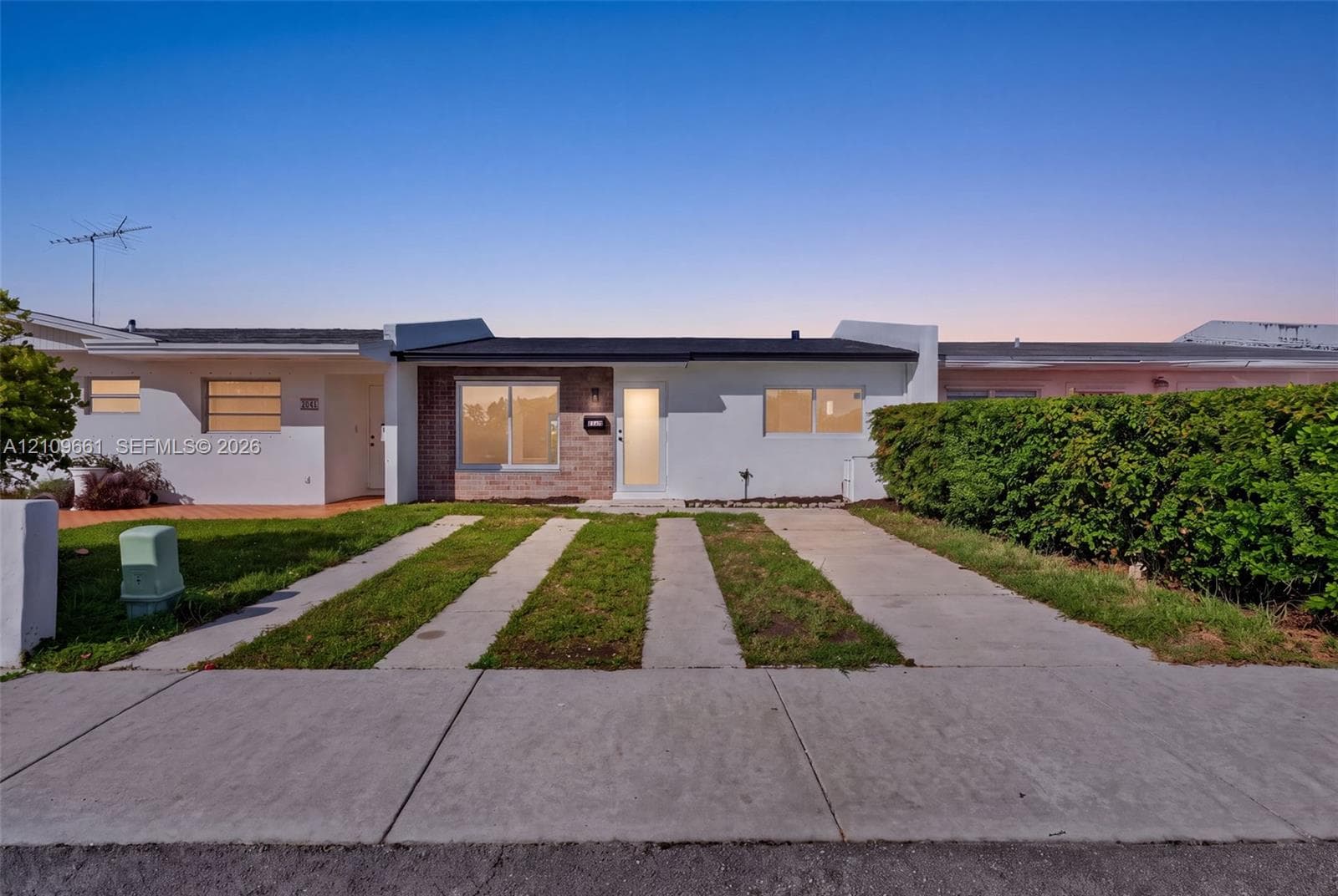 Single-story exterior featuring white stucco siding, stone masonry accents, a concrete ribbon driveway, and a tall privacy hedge