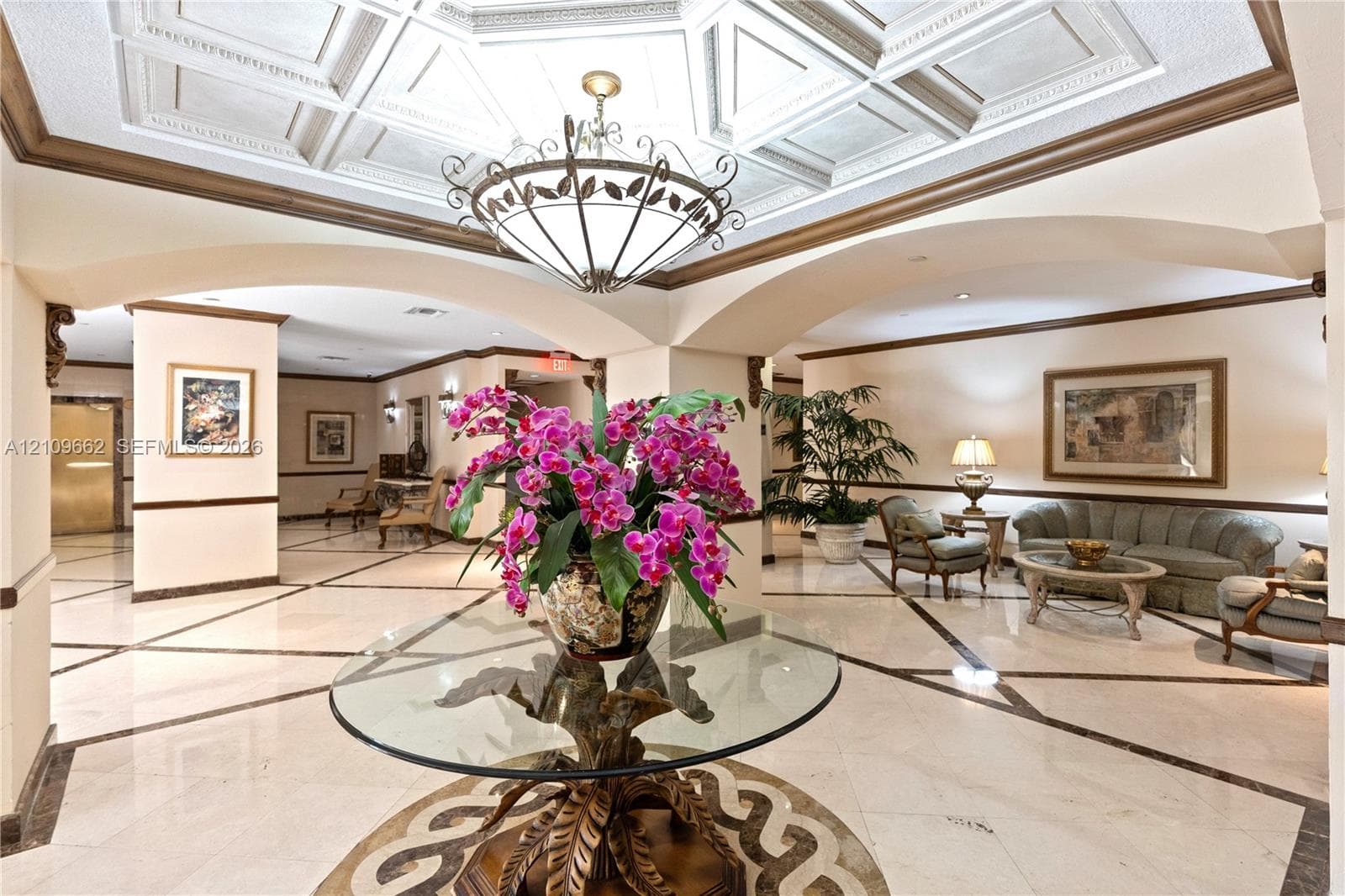 Grand foyer featuring intricate coffered ceiling detailing, stone-finish flooring with decorative inlay, arched architectural openings, and a prominent multi-arm central chandelier