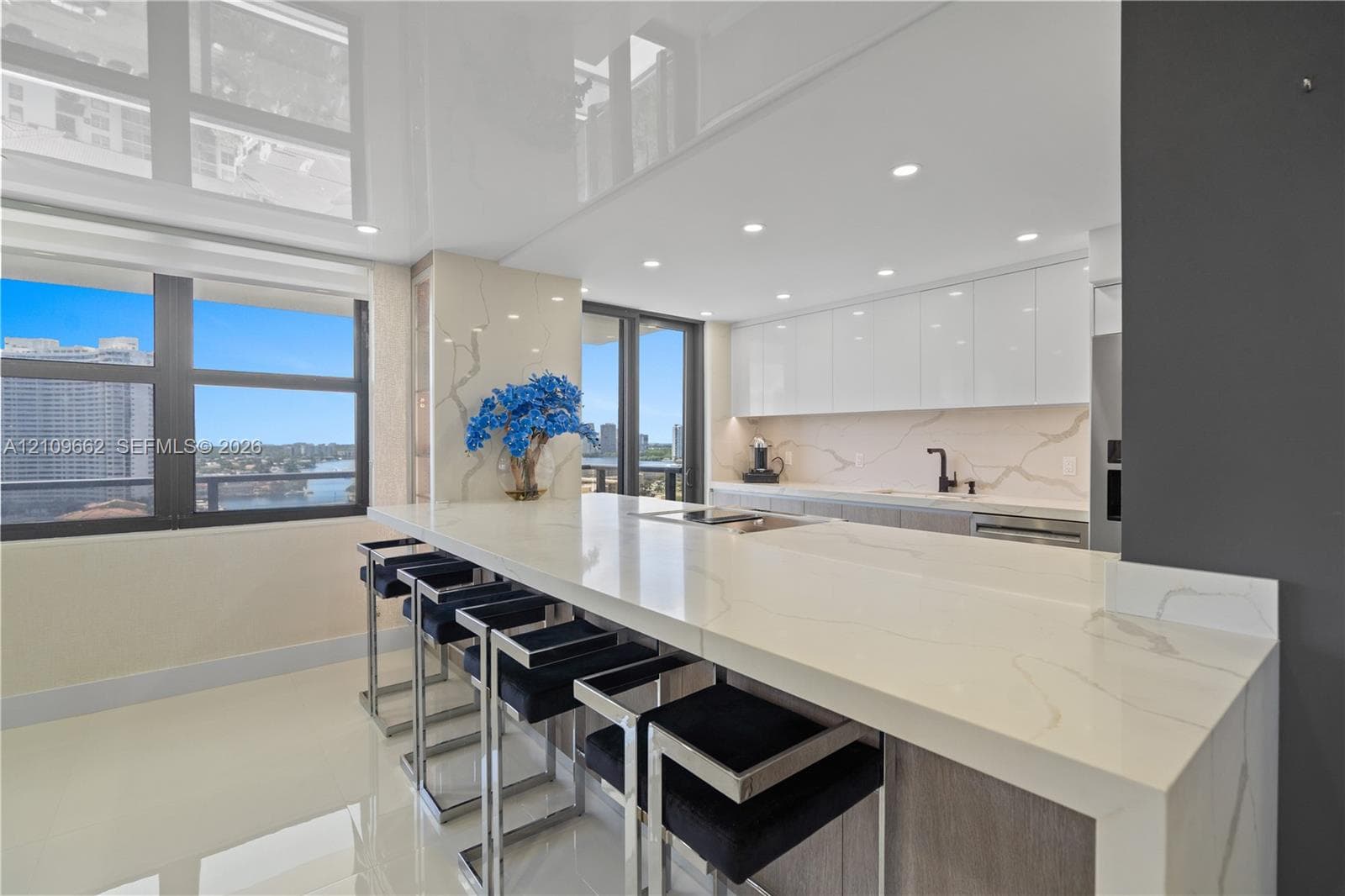Modern kitchen featuring an expansive waterfall-edge stone countertop, high-gloss flat-panel upper cabinetry, full-height stone backsplash, matte black faucet, and recessed ceiling lighting