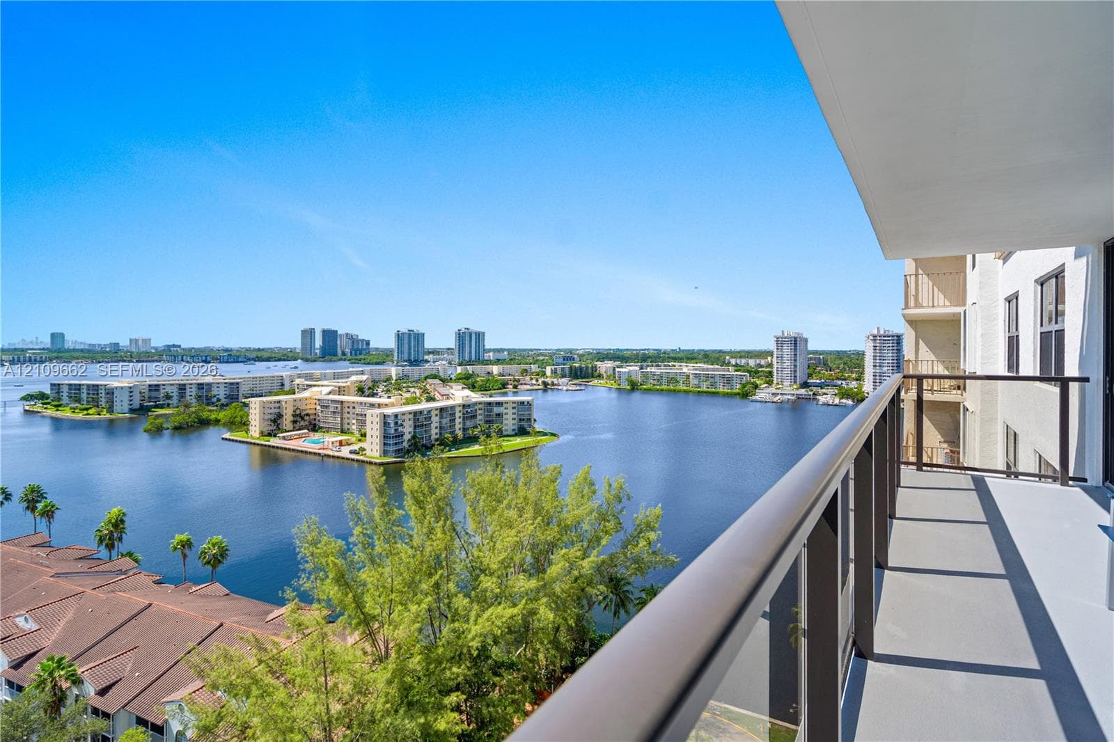 Covered balcony featuring metal railings with glass panels, overlooking a water expanse and surrounding mid-rise structures