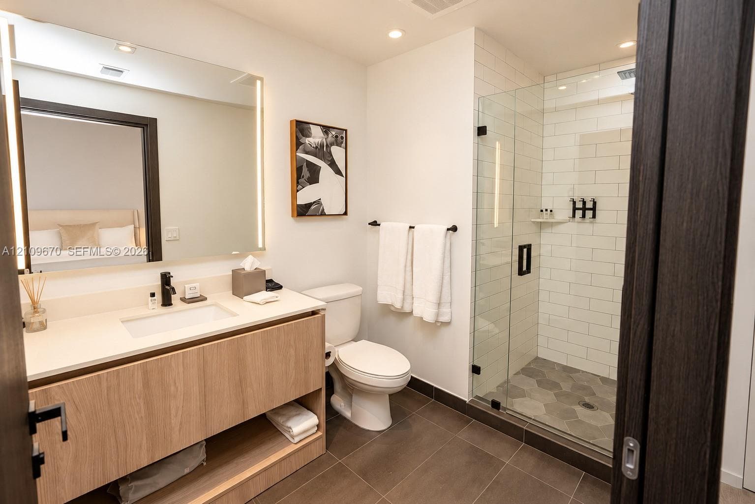 Floating wood-finish vanity with an integrated sink, backlit mirror, glass-enclosed shower with subway tile surround, and hexagonal floor tile