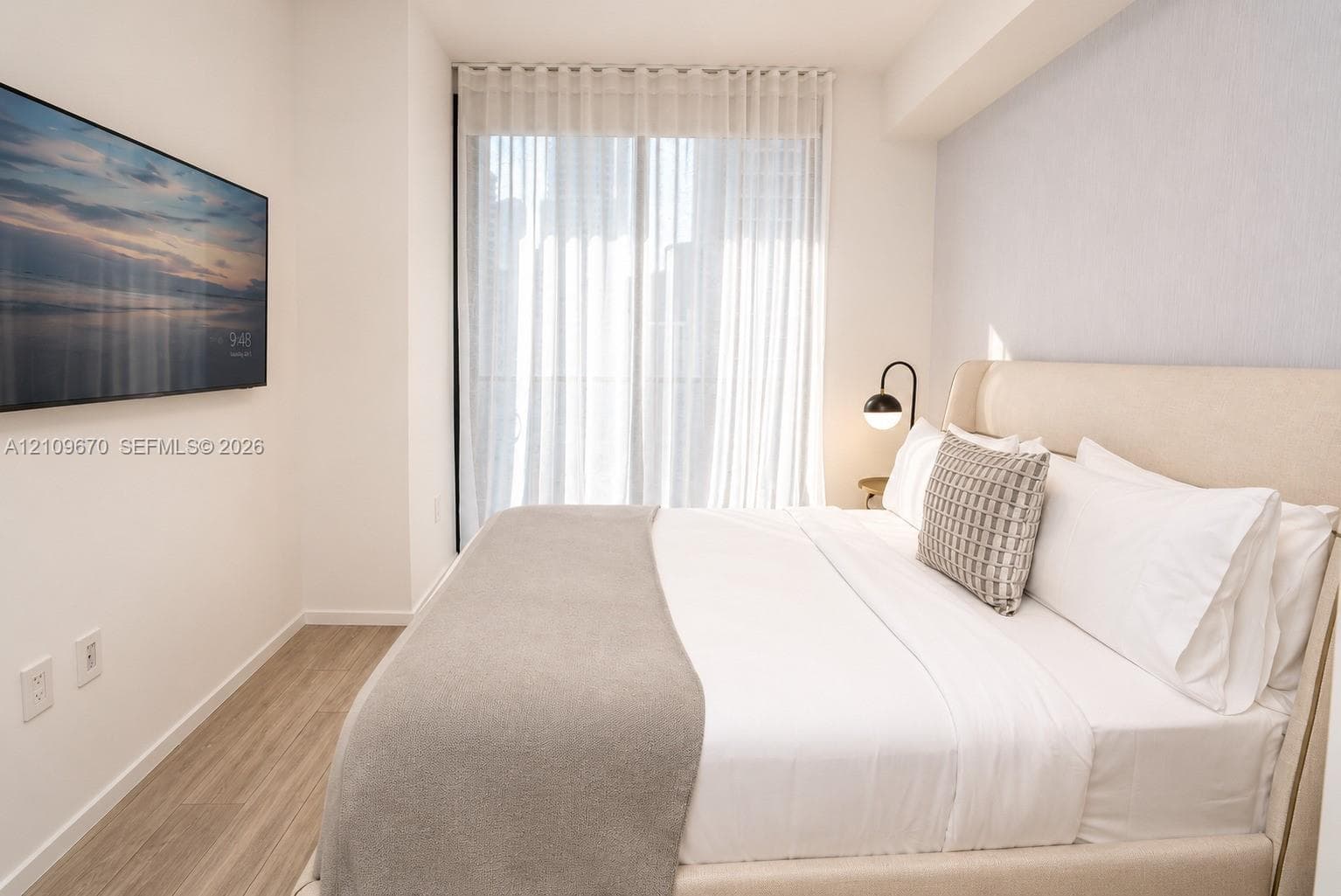 Neutral-toned room featuring wood-finish flooring, a wall-mounted display, floor-to-ceiling drapery, and a minimalist wall-mounted light fixture