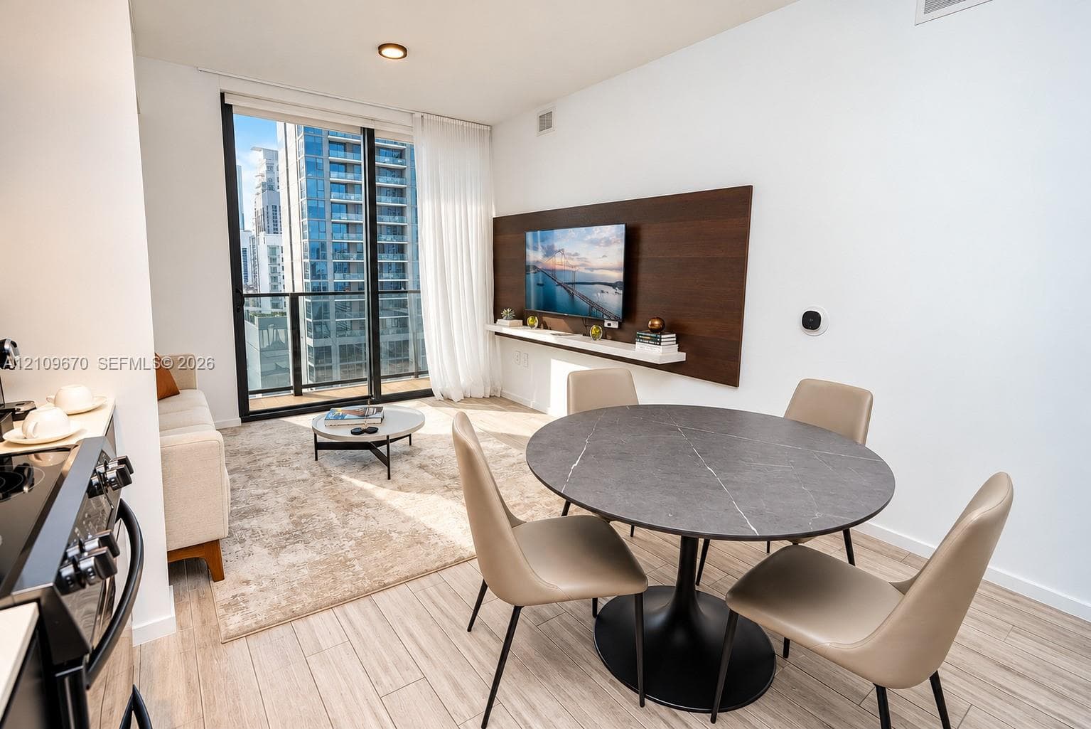Open-concept living space featuring wood-finish flooring, floor-to-ceiling glass sliding doors, a balcony, and a built-in dark wood accent wall with a floating shelf