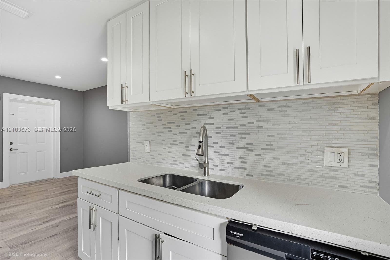 White shaker-style cabinetry, solid surface countertop, stainless steel undermount double-basin sink with gooseneck faucet, horizontal tile backsplash, and wood-finish flooring