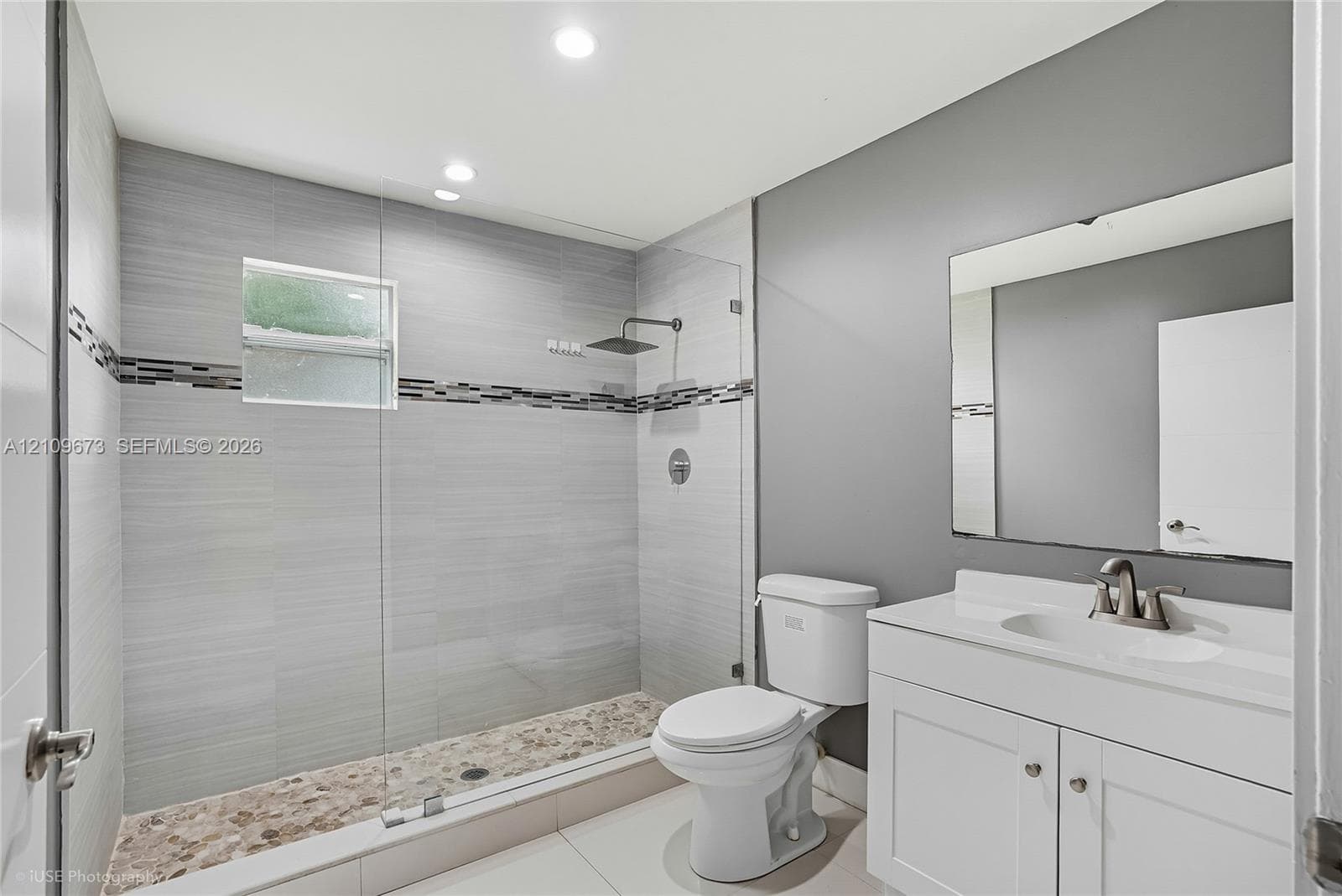 Walk-in shower with floor-to-ceiling gray tile, pebble-stone flooring, frameless glass enclosure, and a rain-style showerhead, complemented by a white vanity with an integrated sink and rectangular wall mirror