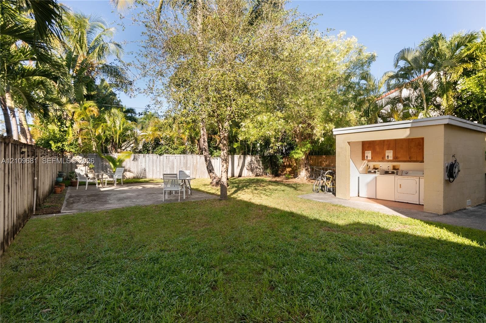 Fenced backyard featuring a manicured lawn, concrete patio, mature trees, and a detached exterior utility structure with built-in cabinetry