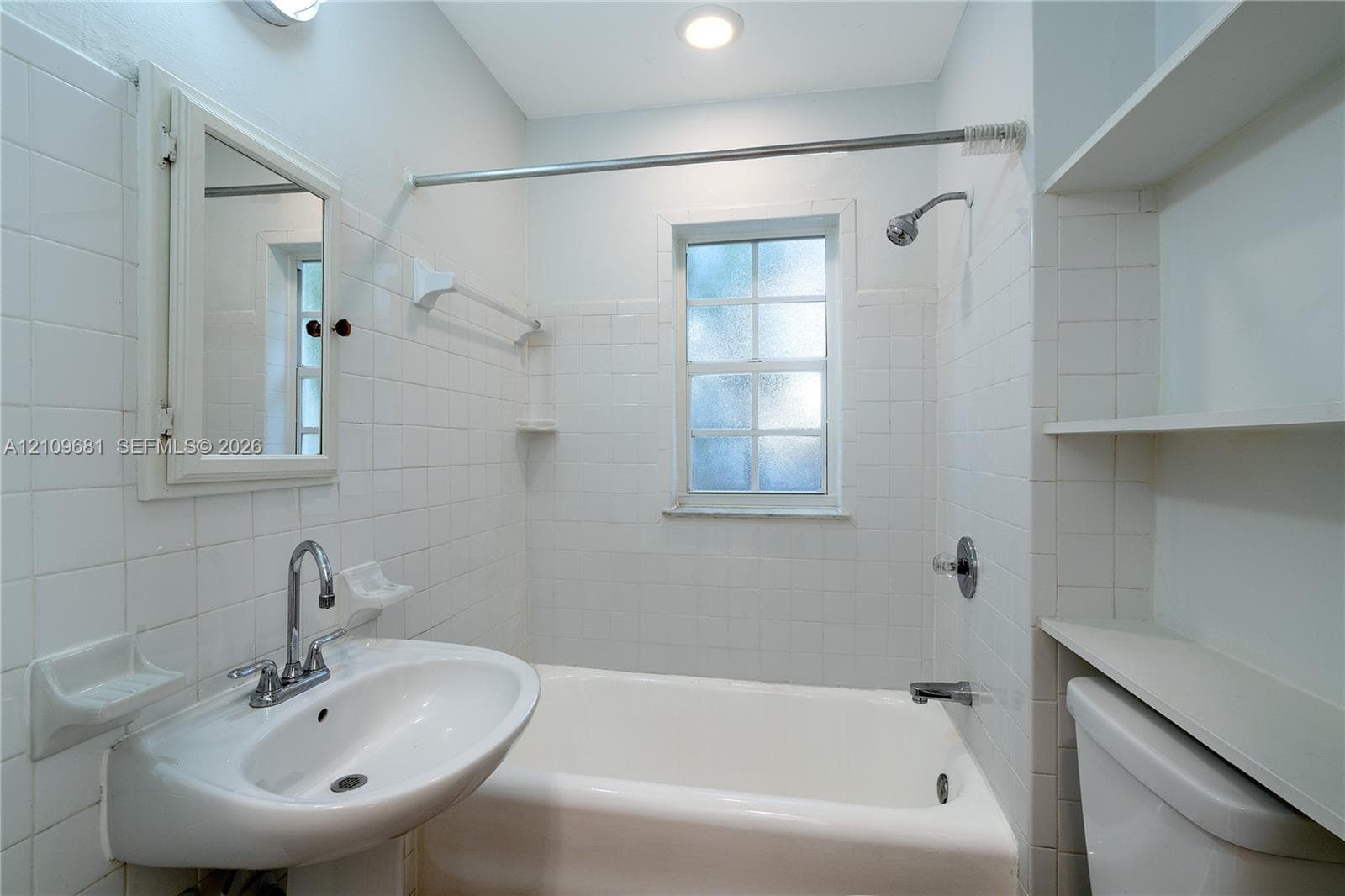 Bathroom featuring white wall tiling, a pedestal sink with a dual-handle faucet, a built-in bathtub with a shower head, integrated wall shelving, and a medicine cabinet