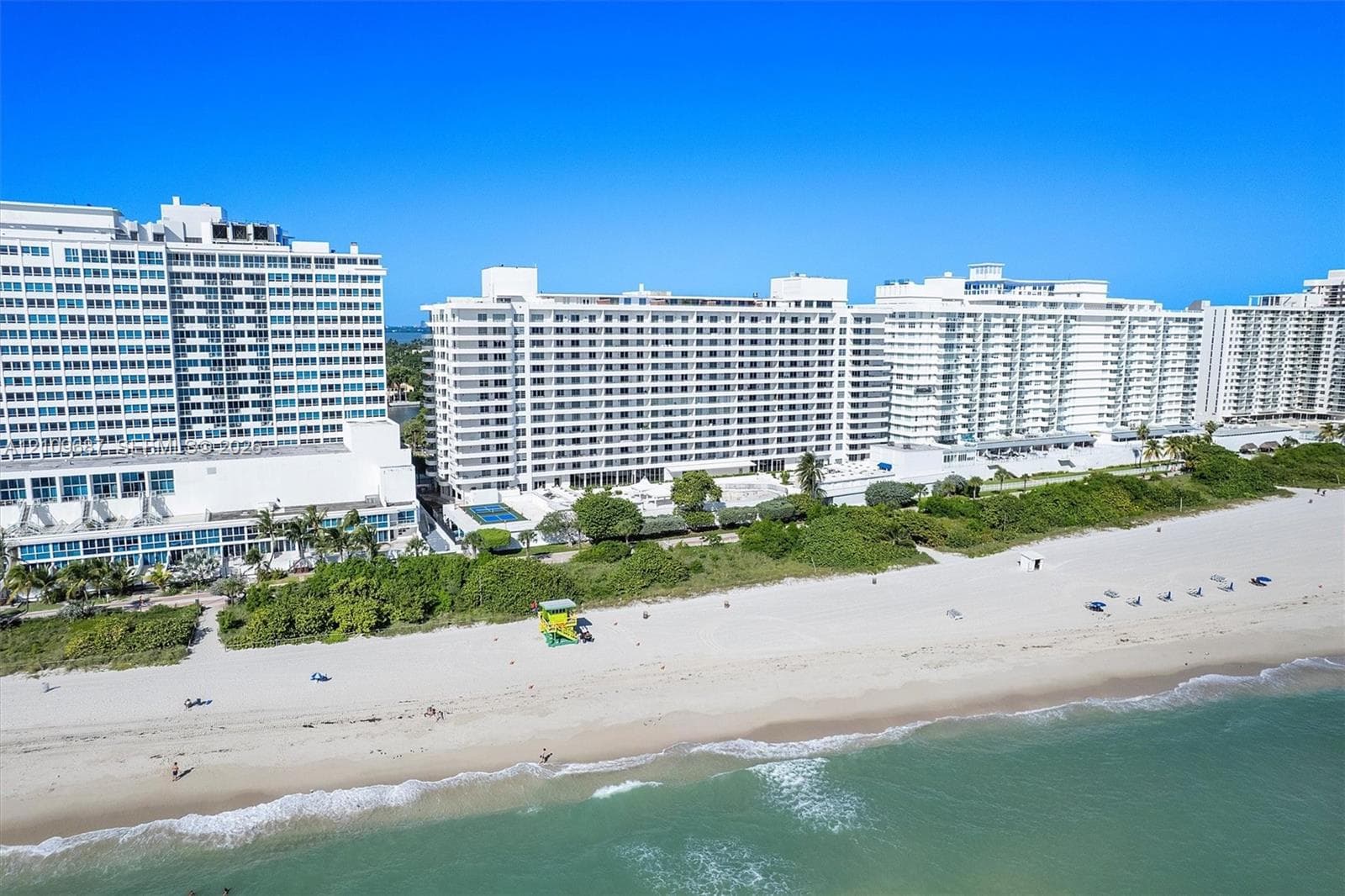 Multi-story beachfront residential buildings featuring white stucco facades, balconies, dense coastal landscaping, and direct access to a sandy shoreline