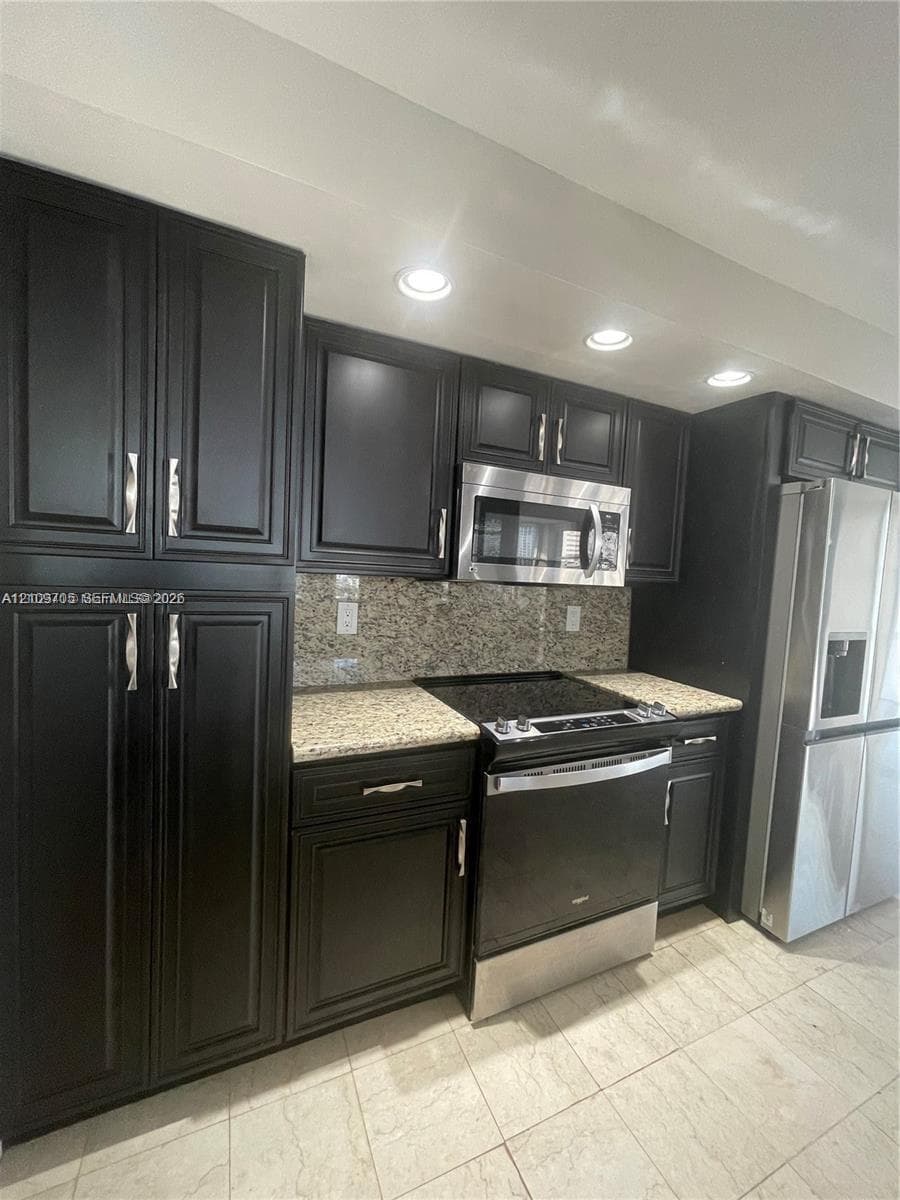 Dark-toned cabinetry featuring shaker-style doors and satin-finish hardware, stone-surface countertops, full-height tile backsplash, stainless steel appliances, and recessed ceiling lighting