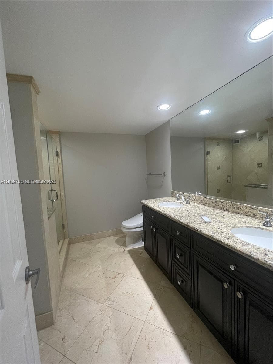 Double vanity with dark cabinetry, light-toned stone countertop, dual undermount sinks, and full-width vanity mirror, featuring tile flooring and a glass-enclosed shower