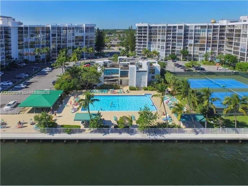 Waterfront complex featuring an outdoor swimming pool, shaded terrace, tennis courts, and mature landscaping