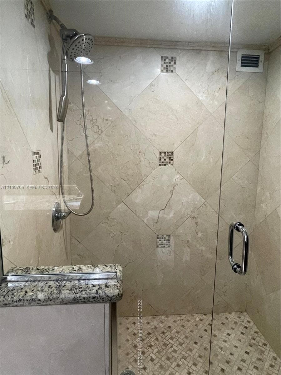 Walk-in shower featuring diagonal-set stone wall tiles, integrated decorative tile inserts, a glass door with a polished metal handle, and a multifunction shower head on a sliding bar