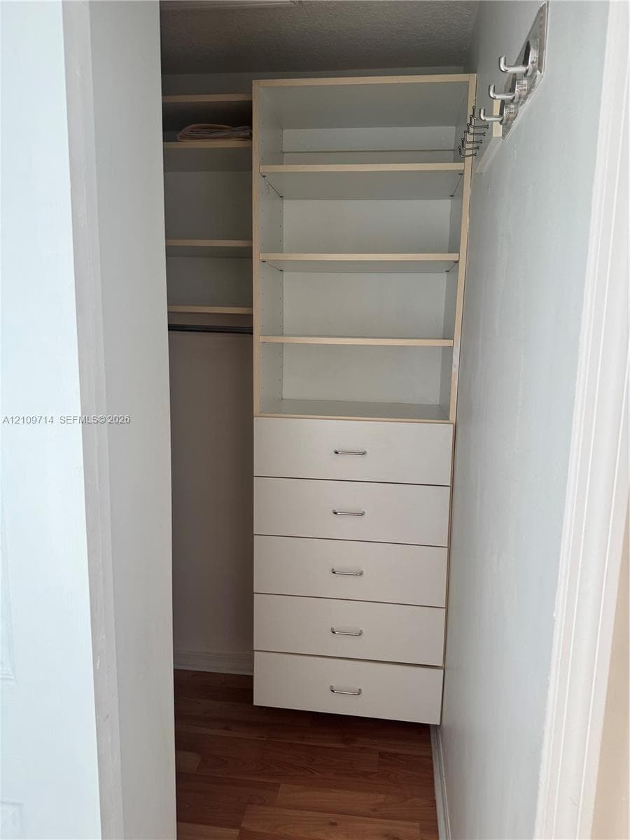 Custom reach-in closet featuring white built-in shelving, a five-drawer dresser unit with metallic pulls, a hanging rod, and wood-finish flooring