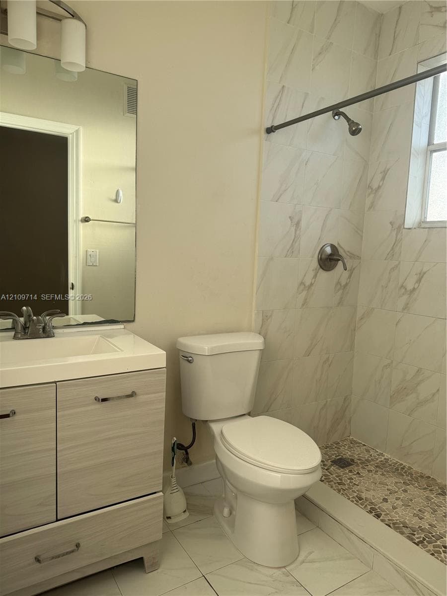 Bathroom featuring a light wood-finish vanity, large wall-mounted mirror, white ceramic sink, shower enclosure with stone-patterned wall tile, and a pebble-style shower floor