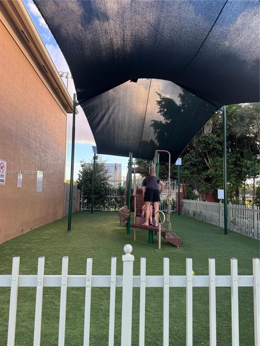Outdoor recreation space featuring synthetic turf, a metal playground structure, and a tension-fabric shade canopy enclosed by white picket fencing