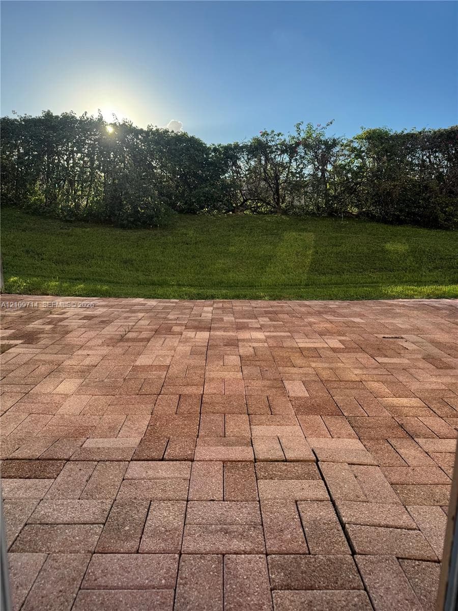 Paver patio featuring a brick-pattern layout, grass lawn, and mature privacy hedge