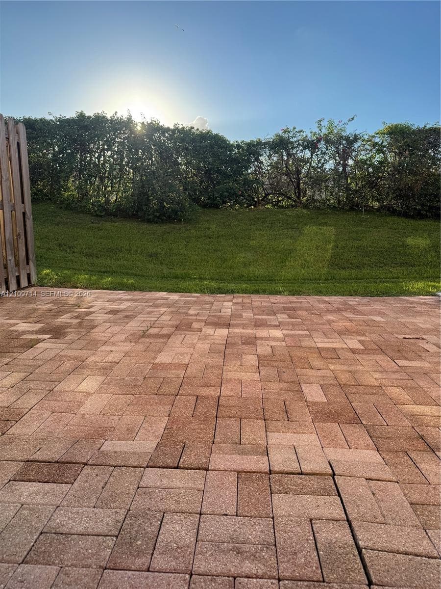 Backyard patio featuring red-toned concrete pavers, an elevated grass lawn, and a perimeter privacy hedge
