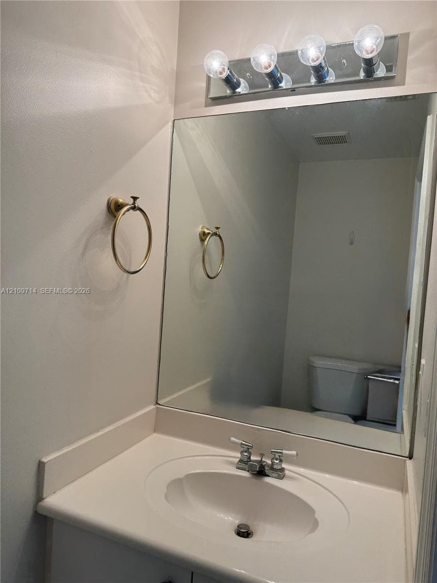 Vanity featuring an oval undermount sink, integrated solid-surface countertop, gold-toned wall-mounted towel ring, four-bulb vanity light fixture, and a wall-to-wall mirror
