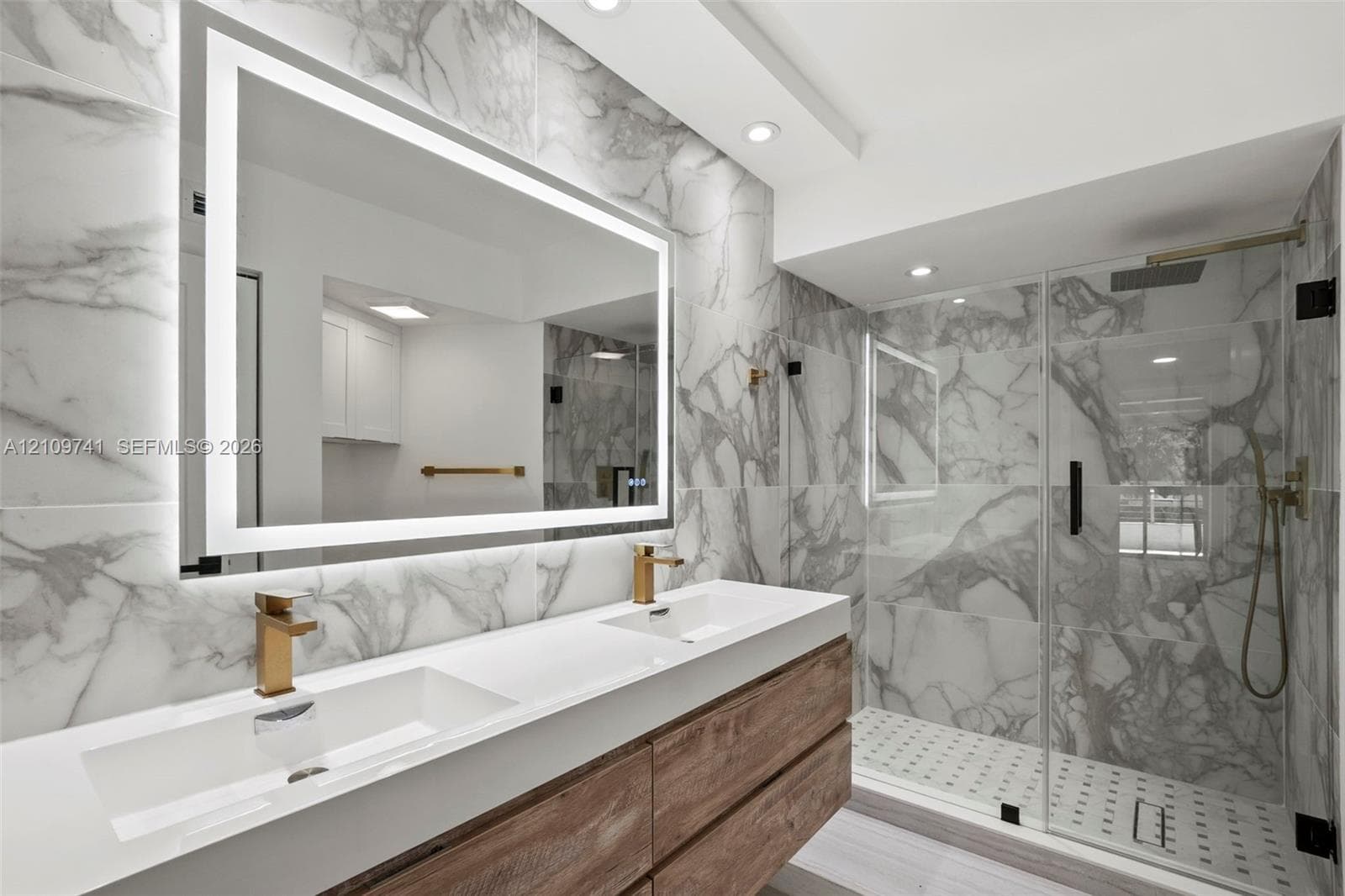 Floating dual-sink vanity with wood-finish cabinetry, backlit LED mirror, large-format stone-patterned tile wall, and glass-enclosed shower with gold-tone hardware