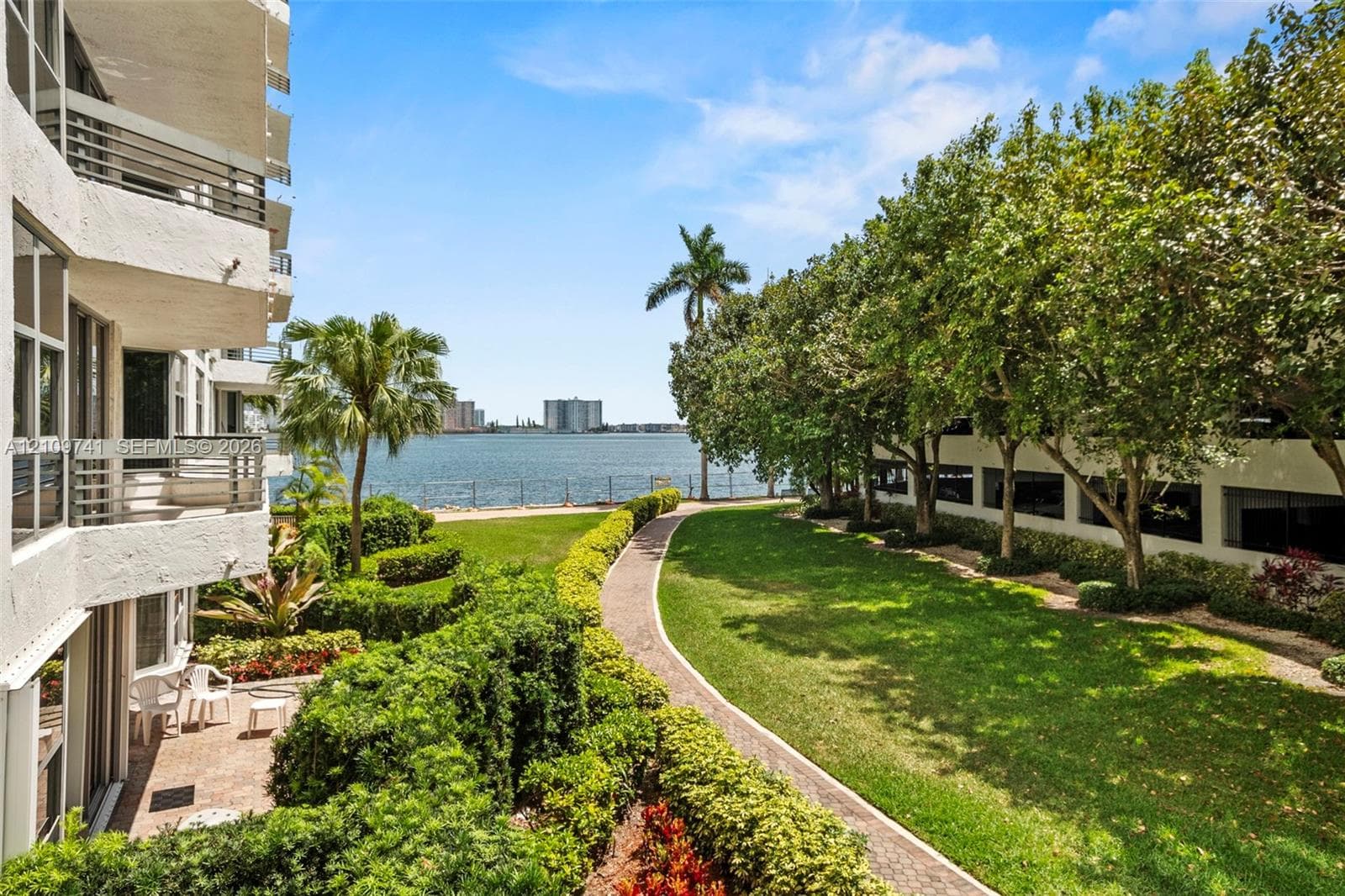 Waterfront grounds featuring a winding stone-paver path, manicured lawn, mature trees, and private balconies with horizontal railings