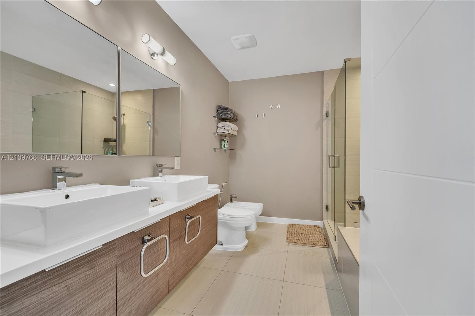 Dual-sink vanity with wood-finish cabinetry, rectangular vessel sinks, large wall-to-wall mirror, glass-enclosed shower, and tile flooring