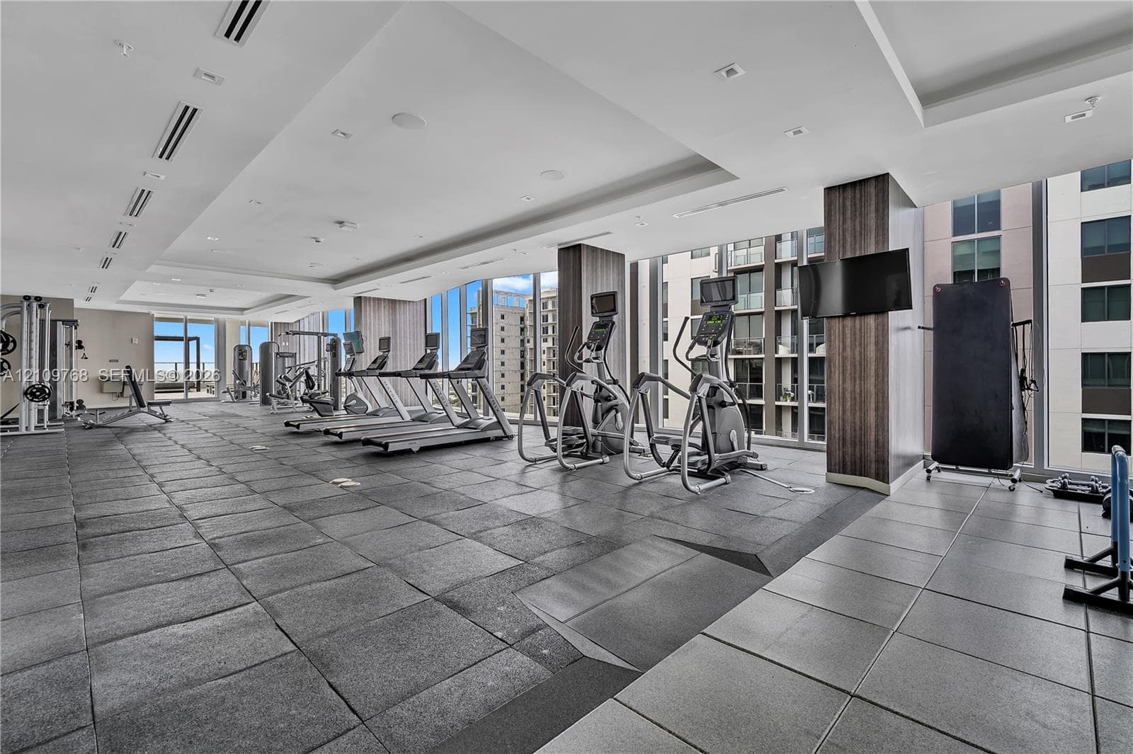 Fitness center featuring floor-to-ceiling windows, tray ceilings with recessed lighting, large-format tile flooring, and wood-finish column detailing
