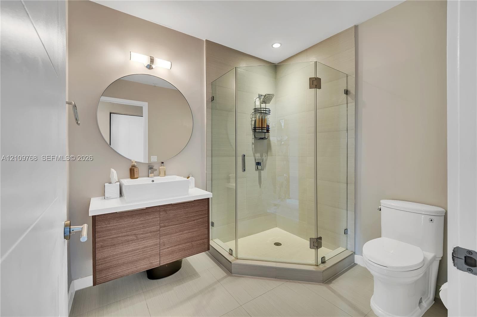 Floating wood-finish vanity with a rectangular vessel sink and white countertop, complemented by a round wall-mounted mirror, brushed-metal light fixture, and a glass-enclosed corner shower with tile surround